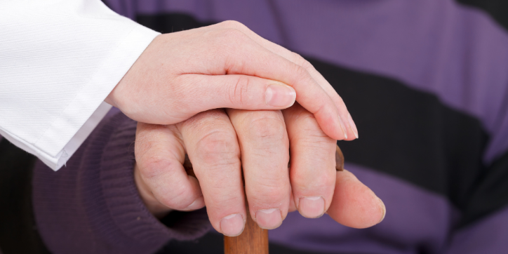 A person wearing white gently rests their hand on top of another person's hand, which is gripping a wooden walking stick. The second person is wearing a purple and black striped jumper.