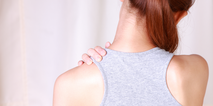 A person with brown hair in a ponytail, wearing a grey vest top, is seen from behind, holding their left hand on their right shoulder, possibly indicating shoulder or neck discomfort.