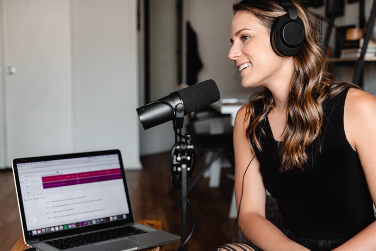 A woman wearing headphones speaks into a microphone whilst recording at a desk with an open laptop displaying text and a chart.