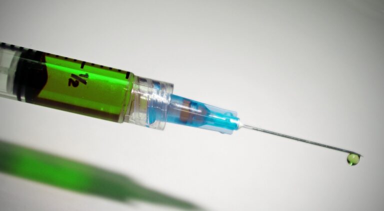 A close-up of a syringe filled with green liquid, with a droplet forming at the tip of the needle against a light background.