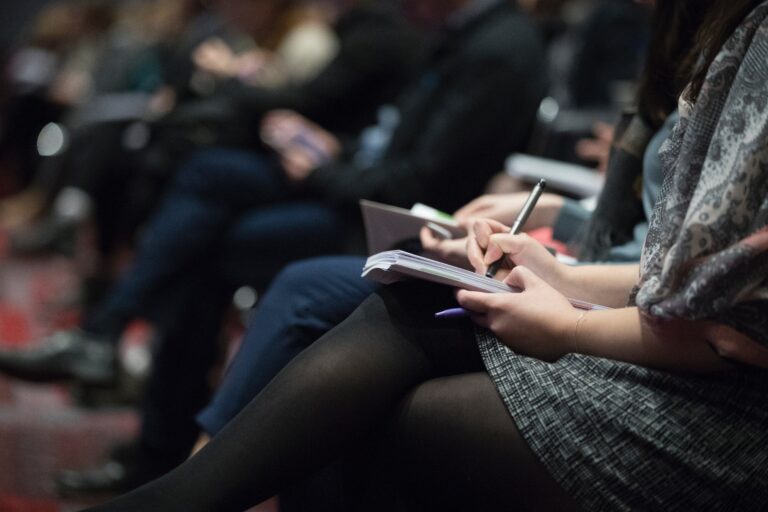 People sitting in rows at an event or conference, with a focus on a person in the foreground taking notes in a notebook whilst others also listen and write.