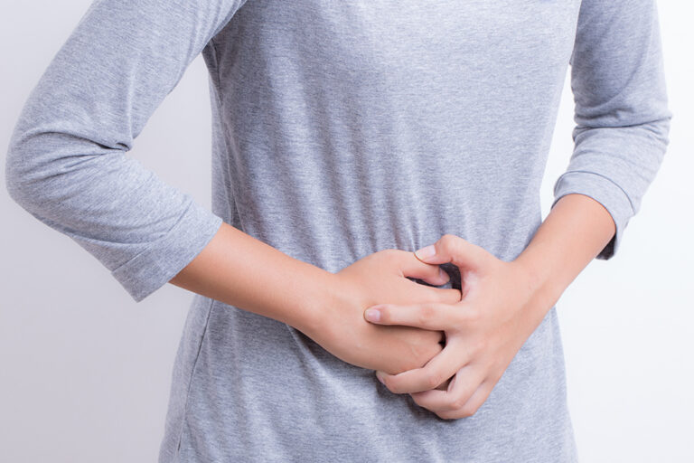 A person wearing a grey long-sleeve shirt holds their stomach with both hands, appearing to be in pain or discomfort, against a plain white background.