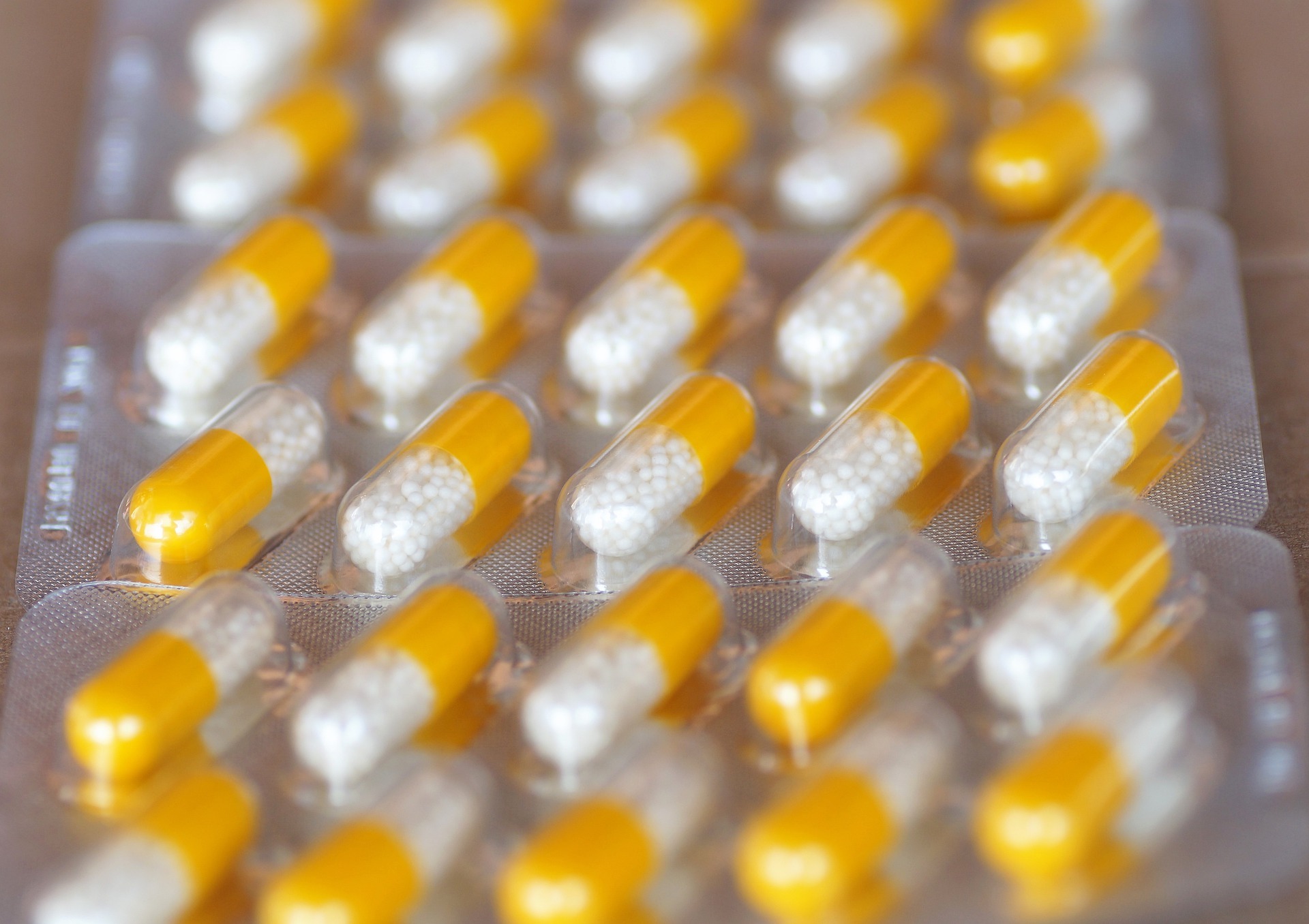 Close-up of several blister packs containing yellow and white capsules, arranged in rows. Each capsule has a yellow end and a clear section filled with small white beads.