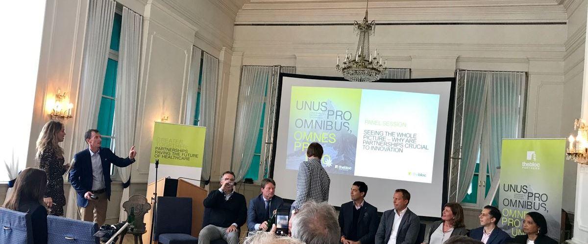A panel discussion is taking place in a bright room. Eight people sit at the front whilst two stand nearby. Two screens display event graphics and text about partnerships and innovation. Audience members sit facing the panel.