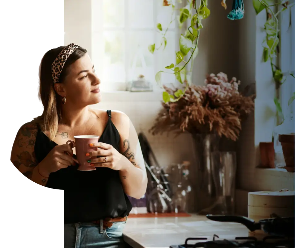 Woman with tattooed arms holding a pink mug, looking out of a sunlit kitchen window surrounded by plants